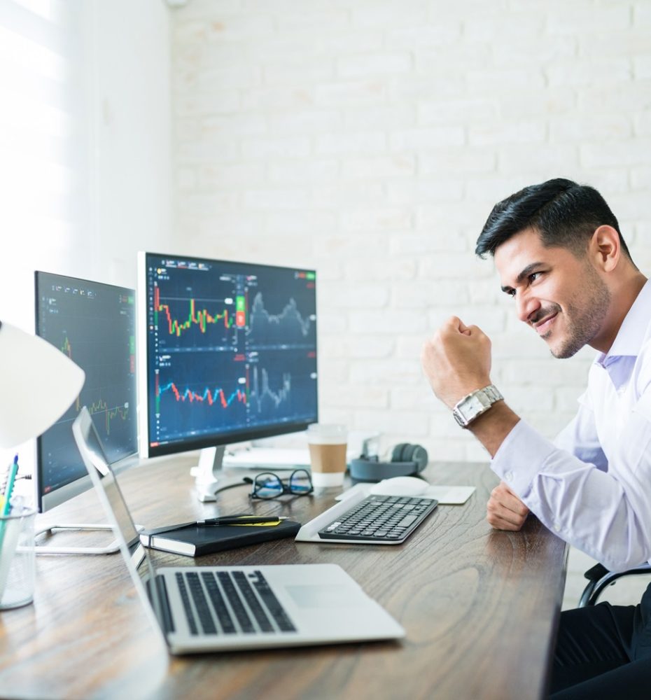 Confident handsome Hispanic financial broker showing fist to computer screen while trading from home